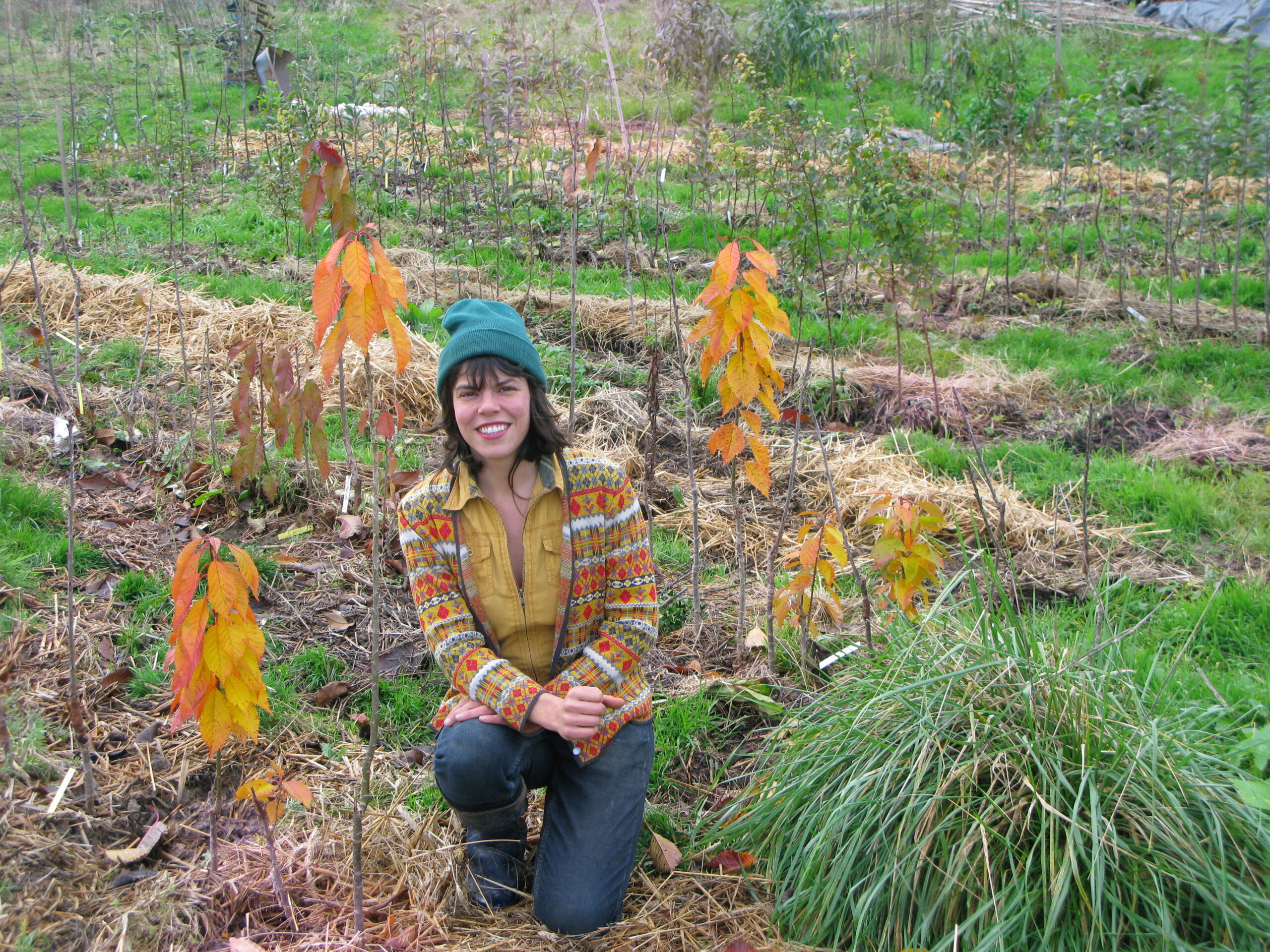 Photo d'une femme accroupie au milieu de jeunes plantations d'arbres. Il s'agit d'un jardin forêt en devenir. Elle porte un bonnet bleu et une veste jaune moutarde avec des motifs. Il s'agit du Jardin Forêt "Les arbres Fruitiers Daumeray" de Laura Bousseronde